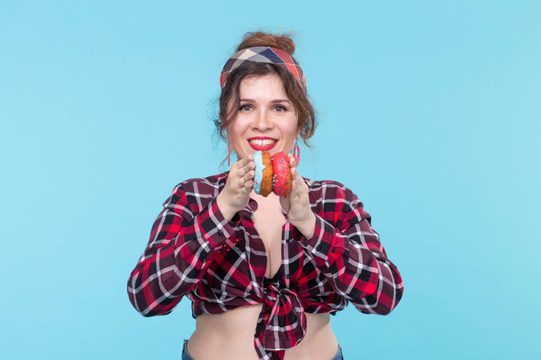 Charming young pin-up girl holding donuts in her hands and one posing on a blue background. Concept benefits and sales of sweets and baking. - Photo, image