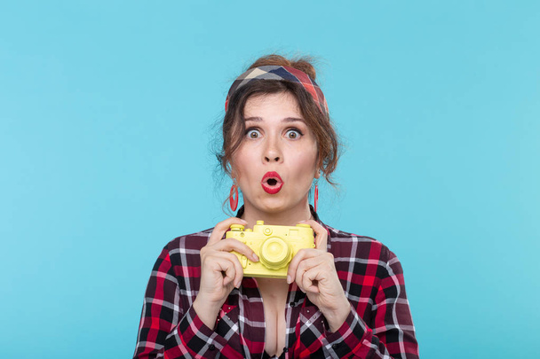 Portret van een mooie jonge positieve vrouw in een geruite shirt houden van een gele film Vintage camera poseren op een blauwe achtergrond. Concept liefhebbers van fotografie en schieten. - Foto, afbeelding