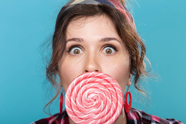 caramelo, dieta y comida chatarra concepto - retrato de mujer pin-up sorprendida con gran piruleta sobre fondo azul
 - Foto, Imagen