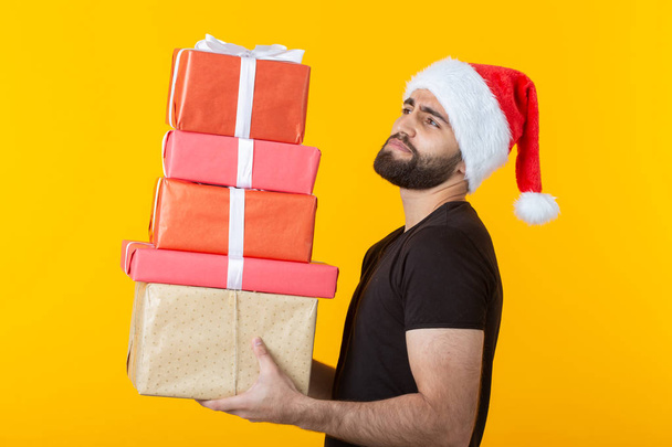 Niezadowolony młody człowiek z brodą w kapeluszu Świętego Mikołaja posiada pięć pudełek upominkowych stwarzających na żółtym tle. Koncepcja prezentów i pozdrowienia dla świąt Bożego Narodzenia i nowego roku. - Zdjęcie, obraz