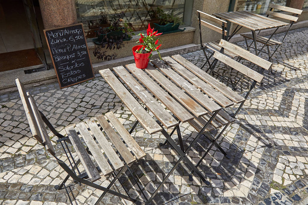 Tavolo e sedie in legno marrone si trovano su un marciapiede portoghese in un caffè all'aperto. Un piccolo vaso di fiori con pepe rosso brillante attira l'attenzione dei passanti. Sullo sfondo c'è un menu del caffè. Sfocatura
. - Foto, immagini
