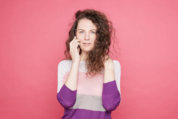 Emotional blue eyed female speaks via cell phone, tells news to friend, dressed in fashionable summer colorful outfit, looks at camera with wonder, stands on pink, copy space aside. Lifestyle concept. - Фото, зображення