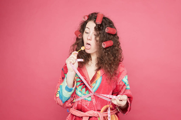 Pretty curly head housewife likes lollipop, remembers taste of childhood, holds candy on stick, dressed in colorful bathrobe, isolated on pink studio wall background. Cheat meal and sweet concept. - Photo, Image