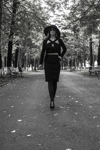 młoda elegancka kobieta w czarnym garniturze biznesowym i czarnym kapeluszu z butelką wody przechadza się latem po słonecznym mieście - Zdjęcie, obraz