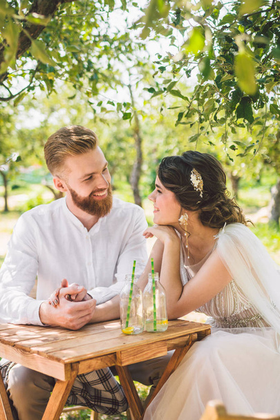 Vonzó pár newlyweds, boldog és örömteli pillanat. menyasszony és vőlegény ül az asztalnál meg a két erdőben. Koncepció romantikus dátum. Esküvői pár ül kávézóasztal és szeretettel nézni egymást - Fotó, kép