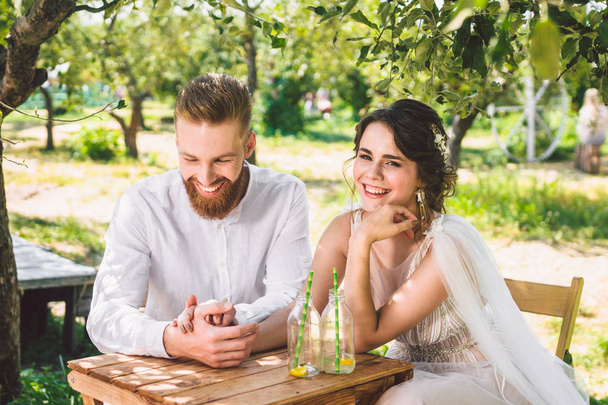 Atraktivní pár novomanželé, šťastný a radostný moment. Nevěsta a ženich sedí u stolu, který je nastavený pro dva v lese. Koncept romantického rande. Svatební dvojice sedí v kavárně a láskyplně se na sebe podívejte - Fotografie, Obrázek