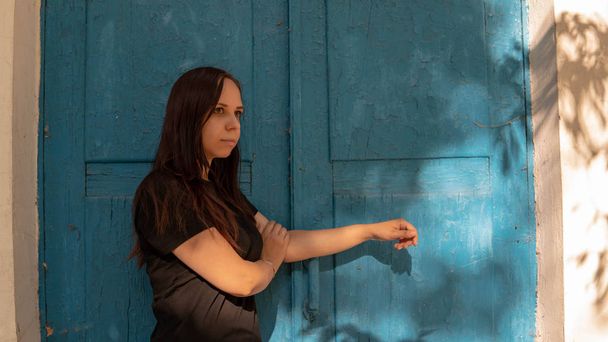 A young woman in a black t-short with long hair stands and looks into the distance on the background of an old building with blue doors. - Фото, изображение