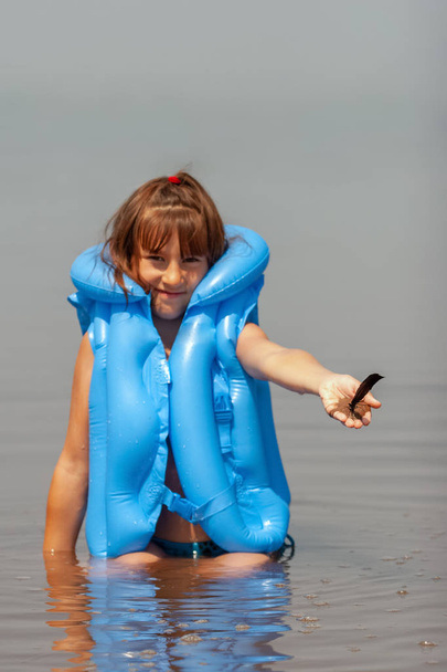 Child girl in a blue inflatable life jacket smiling sits playing in the water and holds out his hand with sand and feather. Selective focus on feather. Copy space. Vertical. - Photo, Image