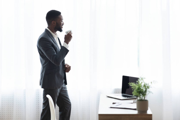 Vrolijke Afro-Amerikaanse zakenman drinken koffie in de buurt van venster in Office - Foto, afbeelding