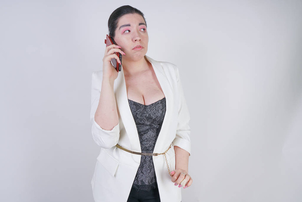 encantadora mezcla de raza más mujer de tamaño en una chaqueta de negocios blanca de pie infeliz y triste con el teléfono inteligente en la mano sobre fondo blanco en Studio
 - Foto, Imagen