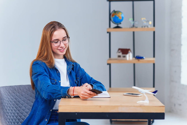 Жінка, одягнена в джинсову сорочку, сидить за столом під час планування відпустки, використовує смартфон
. - Фото, зображення
