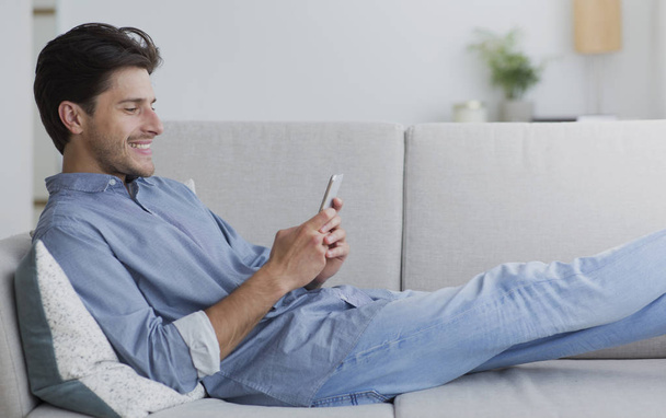 Szczęśliwy człowiek przy pomocy smartphone pasienie się Internet leżący u kanapa - Zdjęcie, obraz