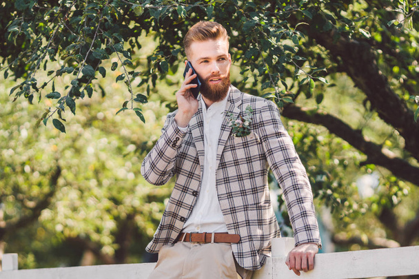 Sexy Brodaty człowiek oczyszczenie z piękną fryzurę z telefonem w ręku. Model ze stylową fryzurą w kratkę i brodę używa telefonu na tle drewna, parku i lasu w słonecznej pogodzie - Zdjęcie, obraz
