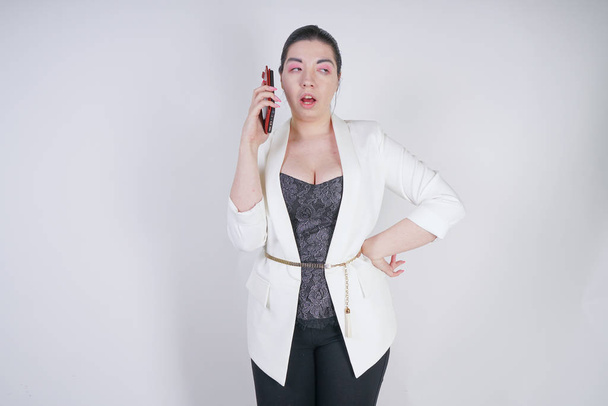 Charmante Mix Race plus Size Frau in weißer Business-Jacke mit Smartphone in der Hand auf weißem Hintergrund im Studio - Foto, Bild