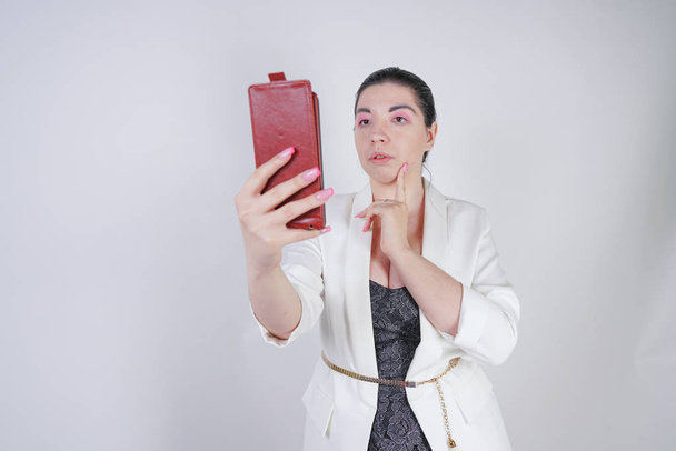 スタジオで白い背景に手にスマートフォンを持って立っている白いビジネスジャケットで魅力的なミックスレースプラスサイズの女性 - 写真・画像