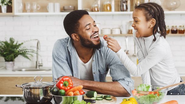 Cute african girl giving her dad cherry tomato while cooking - Photo, Image