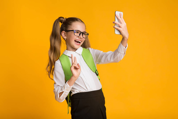 Iskoláslány használ smartphone bevétel Selfie Gesturing V-jel, sárga háttér - Fotó, kép