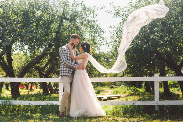 Couple caucasien amoureux mariée et marié debout dans embrasser près de bois blanc, clôture rurale dans le parc d'un verger de pommes. thème est portrait de mariage et belle robe blanche de mariage avec long voile
 - Photo, image