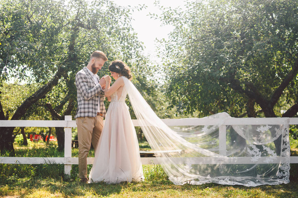Caucasian couple in love bride and groom standing in embrace near wooden white, rural fence in park an apple orchard. theme is wedding portrait and beautiful wedding white dress with long veil - Photo, Image