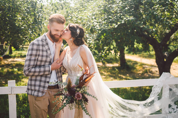 Caucasian couple in love bride and groom standing in embrace near wooden white, rural fence in park an apple orchard. theme is wedding portrait and beautiful wedding white dress with long veil - Foto, Imagen
