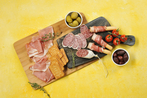 A snack set of slices of salami, bacon, Parma ham. Meat products are supplemented with crackers and grissini breadsticks. Next cups with green and black olives. Yellow background. View from above. - Foto, Bild