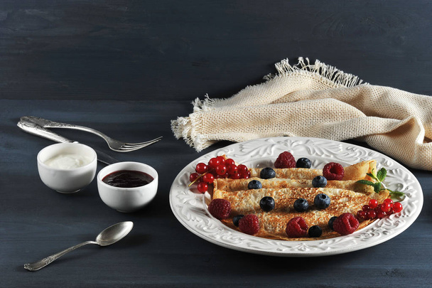 Pancakes on a plate. Pancakes are decorated with berries of red currants, raspberries, blueberries. The frame is completed with a plate with berries, napkins, cutlery. Dark background. Close-up. - Foto, afbeelding