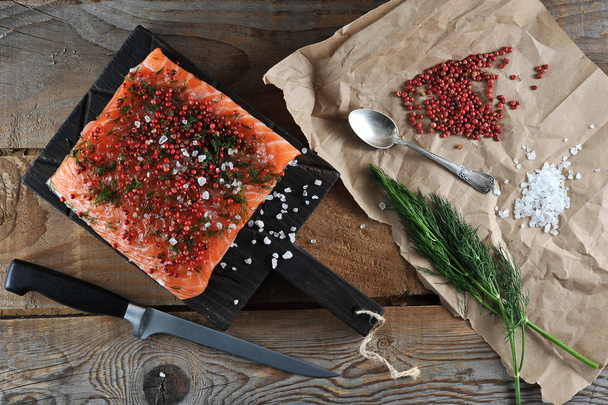 A piece of fresh salmon on a dark board in the process of pickling. The fish is sprinkled with sea salt, pink pepper and dill. Next to kraft paper salt, pepper, dill. View from above. Close-up. - Фото, зображення