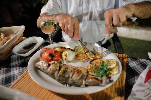 Dinner at the restaurant. Mediterranean food. Grilled fish and vegetables on a plate. Hands of a man cutting fish in a plate. - Photo, Image