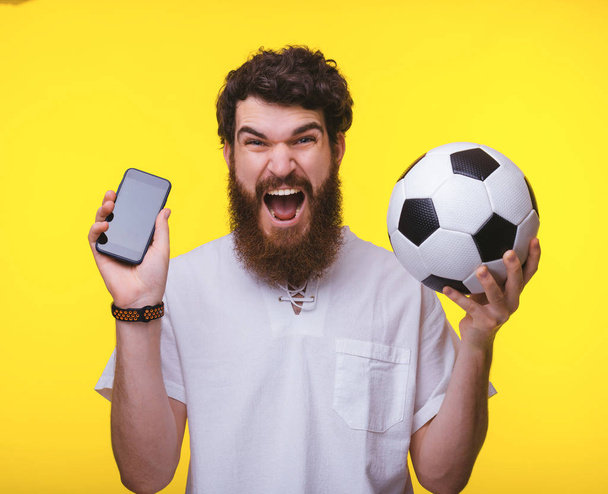 Φωτογραφία του ενθουσιασμένο υποστηρικτή, κρατώντας κινητό και μπάλα ποδοσφαίρου πάνω  - Φωτογραφία, εικόνα