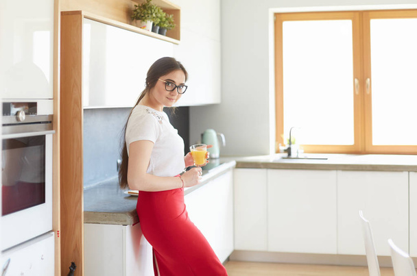 Attractive woman holding a glass of orange juice while standing in the kitchen - Foto, afbeelding