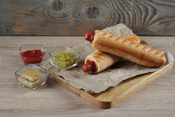 French hot dogs on a wooden board. In cups toppings for hot dog filling: kechchup, mustard, cucumber salad. Light wooden background. - Fotografie, Obrázek