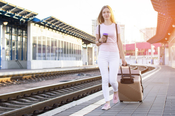attractive girl is standing with luggage at the station and waiting for the train, the student is going on a trip, she is walking along the platform, copy space - Foto, Imagen