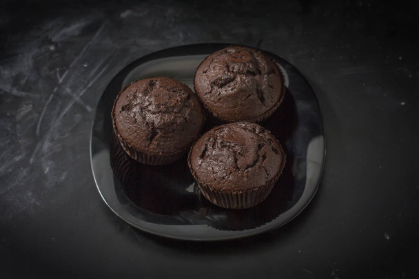 drei Schokoladenmuffins auf einem schwarzen Teller vor dunklem Hintergrund in Nahaufnahme - Foto, Bild