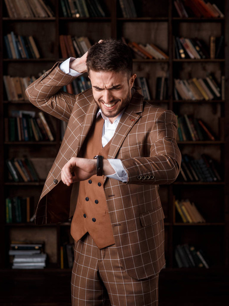 a young man with a beard in a brown suit with a smile sits on his watch and holds his head while standing in the library. A businessman smiles and keeps track of time. - Foto, Imagen