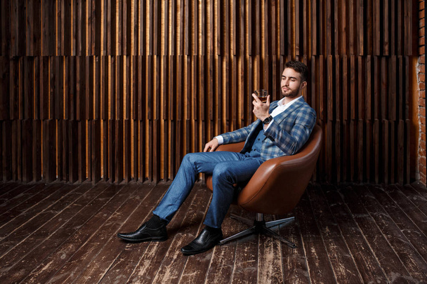 Egy sikeres szakállas üzletember kék kockás öltönyben ül a tetőtéri szoba ablakánál. Egy öltönyös fiatalember whiskyt és konyakot iszik. - Fotó, kép