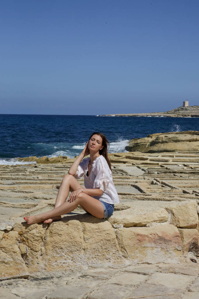 Joven mujer bonita en una camisa de lino blanco y pantalones cortos de mezclilla en la costa rocosa ventosa, disfrutando de vacaciones de verano en la remota ubicación lejos de las atracciones turísticas
 - Foto, imagen