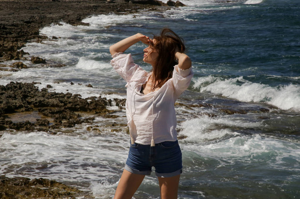 Молода красива жінка в білій лляній сорочці і джинсових шортах на вітряному скелястому узбережжі, насолоджуючись літнім відпочинком на віддаленому місці далеко від туристичних визначних пам'яток
 - Фото, зображення