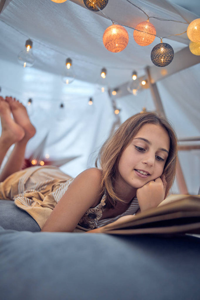10 yaşında küçük bir kız, ev yapımı kitaplarının altında klasik kitaplar okuyor. - Fotoğraf, Görsel