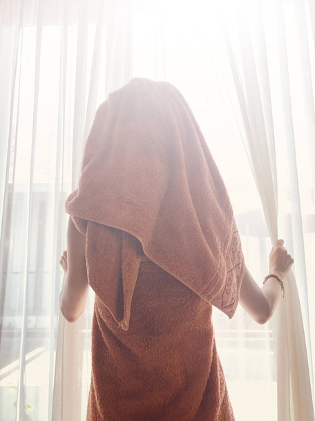 Γυναίκα με πετσέτα μπροστά από ένα παράθυρο και κουρτίνα. - Φωτογραφία, εικόνα