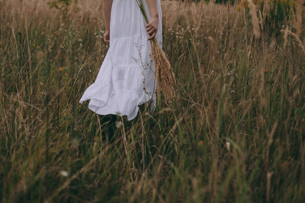 Beyaz elbise ve çizmeler içinde güzel bir genç kadın kırsal yolda yürüyor. Dans ediyor, eğleniyor ve özgürlük yayıyor. - Fotoğraf, Görsel