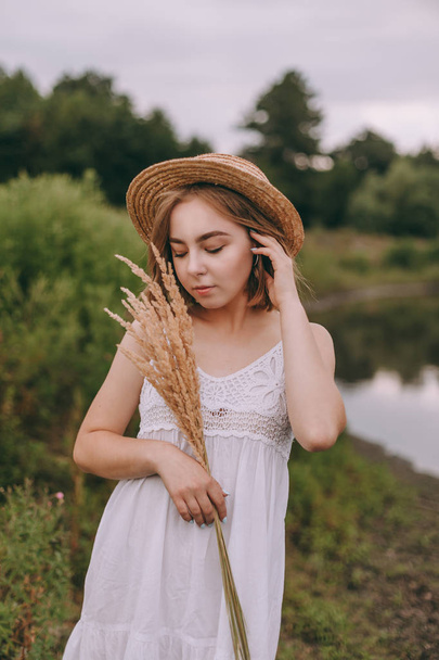 Piękna boho dziewczyna stojąca o zachodzie słońca w pobliżu jeziora. atrakcyjna młoda kobieta w białej cygańskiej sukience z wietrznymi włosami trzymająca słomkowy kapelusz relaksujący w pobliżu wody wśród zielonych liści. letnie wakacje - Zdjęcie, obraz