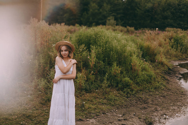 Schöne Boho Mädchen bei Sonnenuntergang in der Nähe des Sees. attraktive junge Frau in weißem Bohème-Kleid mit windigem Haar und Strohhut, die es sich inmitten grüner Blätter am Wasser gemütlich macht. Sommerferien - Foto, Bild