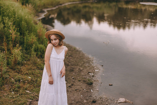 Beautiful boho girl standing at sunset near lake. attractive young woman in white bohemian dress with windy hair holding straw hat relaxing near water among green leaves. summer vacation - Foto, Imagem