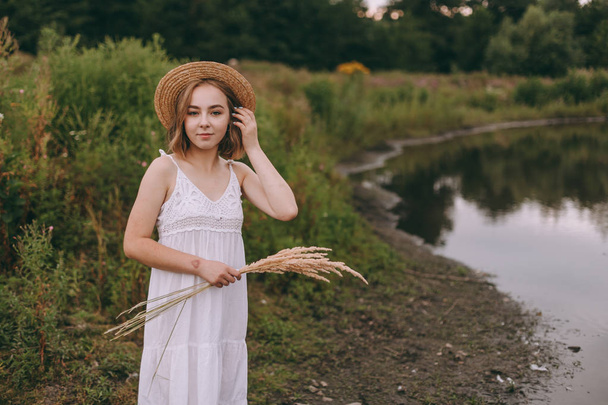 Красивая девушка-бохо, стоящая на закате у озера. привлекательная молодая женщина в белом богемном платье с ветреными волосами, держащая соломенную шляпу, расслабляющуюся у воды среди зеленых листьев. летний отдых
 - Фото, изображение
