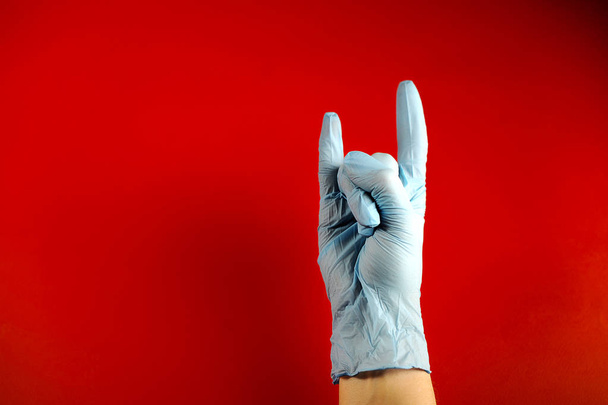 医療用ラテックス手袋に手を入れて人差し指と小指を除いて、すべての指が拳に握り締め付けられます。ジェスチャーは、大胆さや狂気の状態を表現しています。赤い背景。クローズ アップ.  - 写真・画像