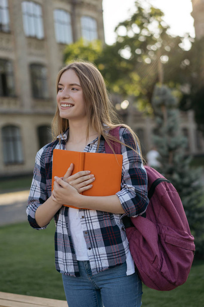 Portrait of happy college girl holding book, standing in park. Successful student with backpack walking to university  - Photo, Image