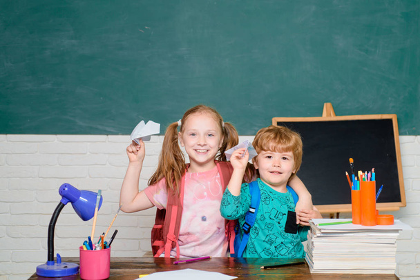 Πίσω στην ιδέα της σχολικής εκπαίδευσης. Παιδιά με χάρτινο αεροπλάνο. Κορίτσι και αγόρι με ευτυχισμένο πρόσωπο έκφραση κοντά στο γραφείο με σχολικές προμήθειες. - Φωτογραφία, εικόνα