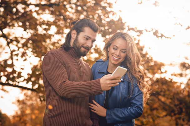 Young couple using cellphone in autumn colored park. - Φωτογραφία, εικόνα
