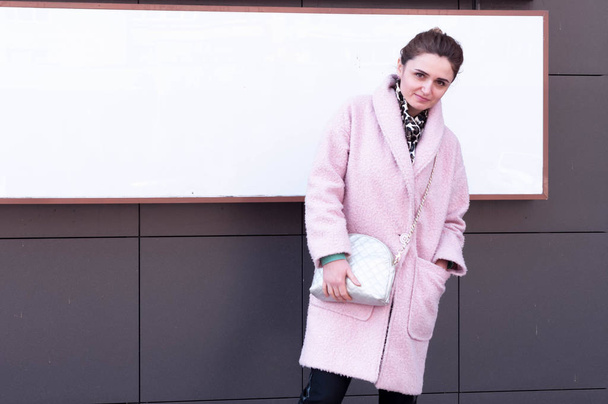 beautiful smiling woman in pink coat and black pants with bundled hair with purse looking at camera in front of blank placard with copy space on sidewalk near building - Fotoğraf, Görsel
