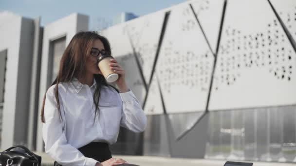 Gyönyörű üzletasszony szemüvegben, okostelefonon beszél és kávét iszik. Modern épület háttérrel - Felvétel, videó